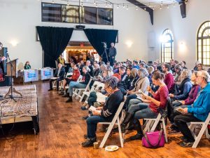 A large room with a crowd of people seated and listening to a person sharing from the stage.