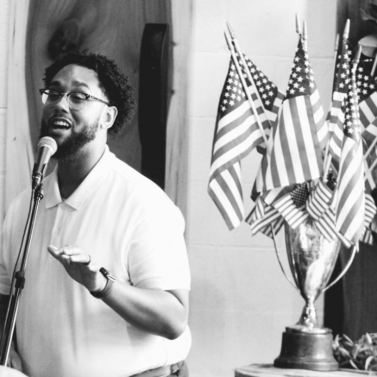 A person speaking into a microphone with American flags in the background.