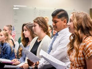 A handful of young people standing together, facing the front of the room, and holding printed pages.
