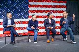 Four people seated and listening to one another on a stage in front of an American flag.