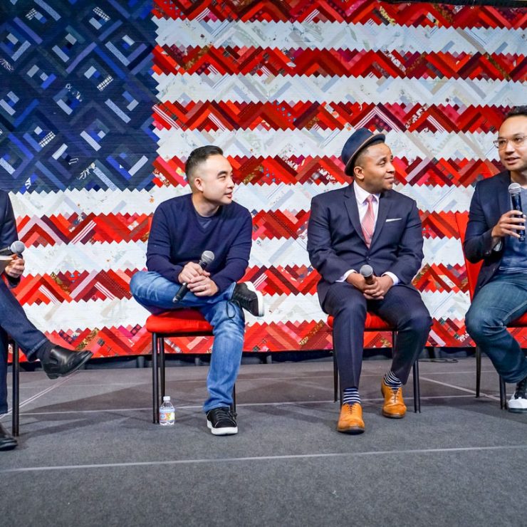 Four people seated and listening to one another on a stage in front of an American flag.