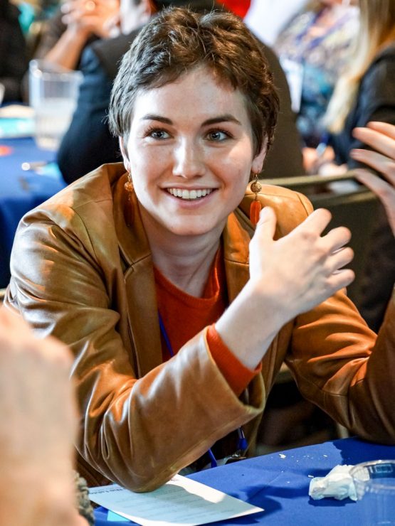A person seated at a table and smiling while talking to others.