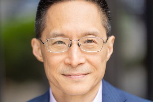 Headshot portrait of an Asian man with a warm, closed mouth smile. He is wearing a pale lilac colored dress shirt under a dark blue twill jacket. He has short black hair with pops of gray, brown eyes and silver rounded corner rectangular glasses, slight stubble above his upper lip, and the background is a soft mix of green and gray tones.