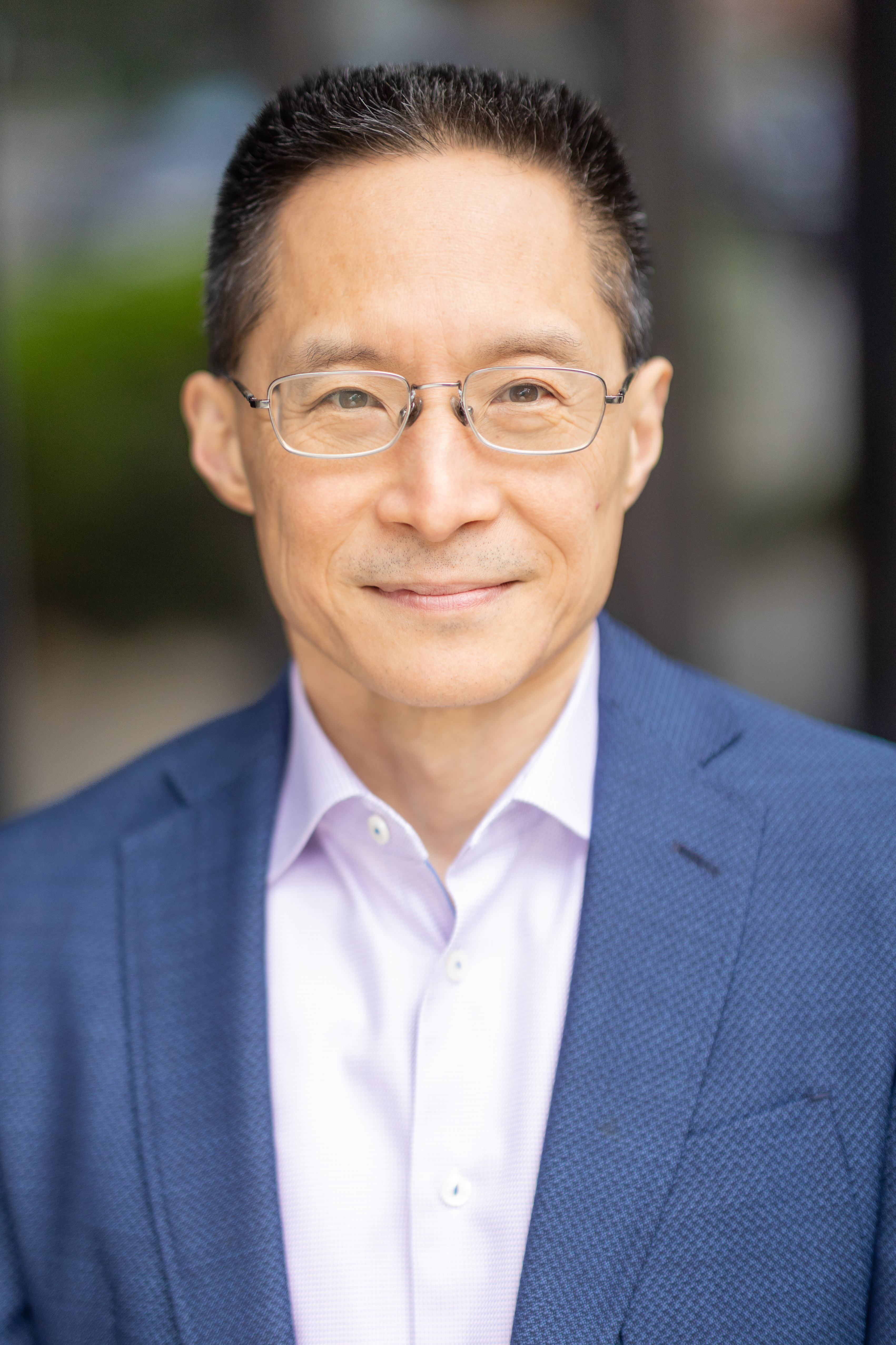 Headshot portrait of an Asian man with a warm, closed mouth smile. He is wearing a pale lilac colored dress shirt under a dark blue twill jacket. He has short black hair with pops of gray, brown eyes and silver rounded corner rectangular glasses, slight stubble above his upper lip, and the background is a soft mix of green and gray tones.